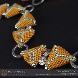 Op de afbeelding: Een close-up van een gouden en zilveren kralenketting met een driehoekig patroon. De ketting is gemaakt van kleine, vierkante kralen die samen geweven zijn om een geometrisch ontwerp te cre&euml;ren. De kralen zijn warm goudkleurig met zilveren accenten.