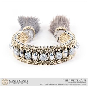 Peut inclure: Un bracelet en perles dor&eacute;es et argent&eacute;es avec un pompon en fourrure grise &agrave; chaque extr&eacute;mit&eacute;. Le bracelet s'appelle "The Tudor Cuff" et est un design original de Manek Lady.