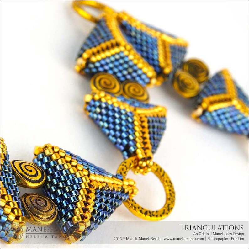 Op de afbeelding: Een close-up van een kralenketting met een geometrisch patroon. De ketting is gemaakt van blauwe en gouden kralen en heeft een driehoekige vorm. De tekst "TRIANGULATIONS" is zichtbaar in de afbeelding.