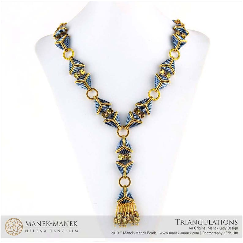 Op de afbeelding: Een blauwe en gouden kralenketting met een driehoekige hanger. De ketting is gemaakt van kleine kralen en heeft een geometrisch ontwerp. De hanger is een grote driehoek met een kleinere driehoek erin. De ketting ligt op een witte achtergrond.
