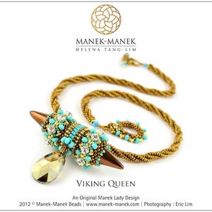May include: A gold and turquoise beaded necklace with a large, ornate pendant featuring a teardrop-shaped crystal. The pendant is shaped like a spearhead and has a gold metal tip. The necklace is called "Viking Queen" and is an original Manek Lady design.