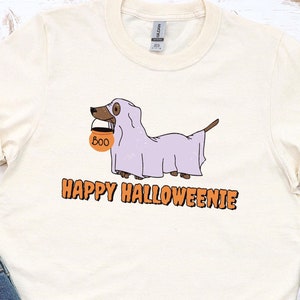 May include: A white t-shirt with a cartoon image of a dachshund dressed as a ghost holding a trick-or-treat bag that says "BOO". The text "Happy Halloweenie" is printed below the image.