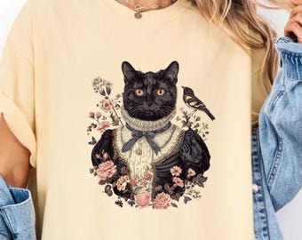 Op de afbeelding: Een lichtgeel T-shirt met een zwarte kat die een witte kanten kraag en een zwarte jurk draagt. De kat wordt omringd door roze en witte bloemen en een kleine vogel.