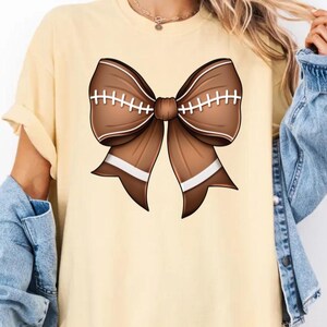 May include: A light yellow t-shirt with a brown football-themed bow on the front. The bow has white stitching and white stripes.