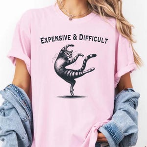 May include: A pink t-shirt featuring a black and white illustration of a cat dancing on pointe shoes. The text "EXPENSIVE & DIFFICULT" is printed above the cat.