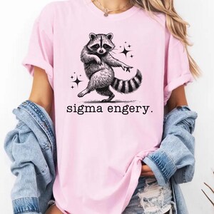 May include: A pink t-shirt with a black and white illustration of a raccoon dancing. The text "sigma engery." is printed below the raccoon.
