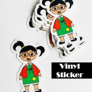 May include: A stack of vinyl stickers featuring a cartoon girl with black hair, glasses, and a red jacket. The girl is wearing a green dress with a yellow pocket and white socks. The text "Vinyl Sticker" is printed below the stickers.