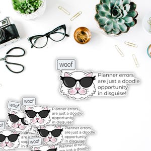 May include: White cat sticker wearing sunglasses with the word "woof" in a speech bubble. Text on the sticker reads "Planner errors are just a doodle opportunity in disguise!" Stationery items and plants are visible.