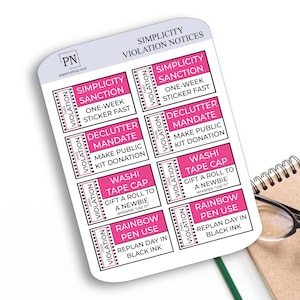 May include: A sheet of white planner stickers with pink and black text, including "Simplicity Sanction" and "Declutter Mandate." A small notebook and glasses are in the background.