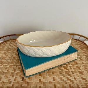 May include: Cream-colored oval serving bowl with a woven basket design and gold trim. The bowl sits on a teal book, placed on a woven bamboo tray. The bowl's rim has a scalloped edge.