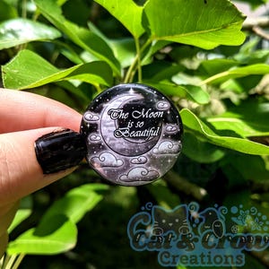 Può includere: Un bottone rotondo nero con un design a luna crescente, nuvole e stelle. Il testo "The Moon is so Beautiful" è presente sul bottone. Il bottone è tenuto davanti a uno sfondo di foglie verdi.