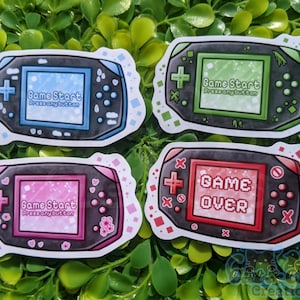 May include: Four different designs of stickers featuring a handheld video game console. The console is black with a colorful screen. The screens display the text "Game Start Press any button" and "Game Over".