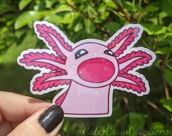 May include: A pink axolotl sticker with a holographic finish. The sticker features a cartoon axolotl with pink gills and a wide open mouth. The sticker is held in front of a green leafy background. The text "Cat&Crow Creations" is at the bottom.