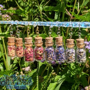 May include: A collection of small glass bottle earrings filled with dried flowers and herbs. Each bottle is sealed with a cork and hangs from a silver or gold-colored hook. The earrings are displayed against a backdrop of green foliage.