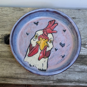 May include: A round ceramic plate with a glazed finish featuring a hand-painted illustration of a white chicken head with a red comb and yellow beak. The background is a mottled pink and purple hue, decorated with several small black heart shapes.