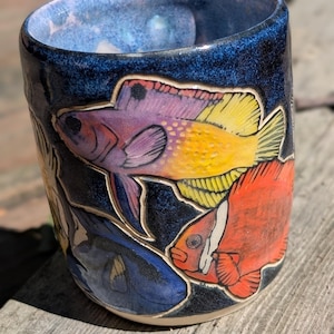 May include: A ceramic mug with a dark blue speckled glaze featuring hand-painted fish in vibrant colors. The design includes a purple and yellow fish, a bright orange fish, and a dark blue fish, all rendered with detailed outlines on the exterior.