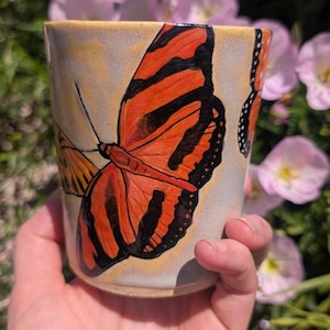 May include: A cylindrical ceramic mug featuring a hand-painted design of vibrant orange and black monarch butterflies against a light, cream-coloured background. The artwork wraps around the exterior surface, showcasing detailed wing patterns.