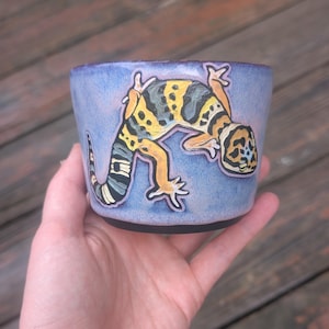May include: A ceramic cup with a light purple glaze featuring a hand-painted leopard gecko. The gecko is depicted in shades of yellow, black, and white with a curved body shape. The cup has a dark base and a smooth, rounded rim design.