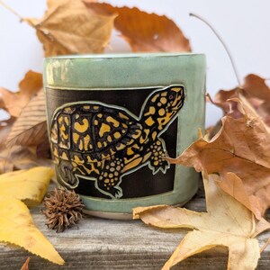 May include: A cylindrical ceramic mug with a light green glaze featuring a rectangular black panel. Inside the panel is a relief carving of a turtle with a yellow and black patterned shell and skin, depicted in profile facing right.