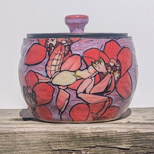 May include: A round ceramic jar with a matching lid featuring a hand-painted design of a pink orchid mantis resting among large red orchid flowers. The background is a textured purple glaze, and the lid has a small, rounded knob handle on top.