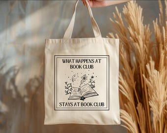 May include: A white canvas tote bag with black text that reads "What happens at book club stays at book club". The text is surrounded by a black border and a black illustration of an open book with flowers and a crescent moon.