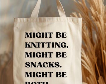 May include: Cream-colored tote bag with black text that reads "MIGHT BE KNITTING, MIGHT BE SNACKS, MIGHT BE BOTH." The bag has two handles and is held up by a hand. The background includes dried ornamental grass.