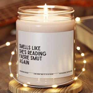 May include: A white candle with a black label that reads "Smells Like She's Reading Faerie Smut Again". The candle is in a clear glass jar with a wooden lid. The candle is surrounded by white string lights.