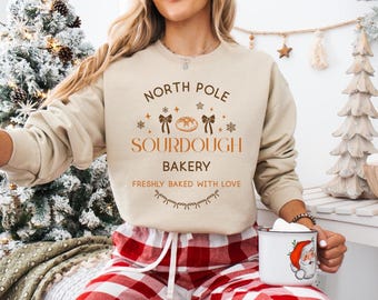 May include: A beige sweatshirt with the text "North Pole Sourdough Bakery" in brown. The design includes illustrations of bread, bows, and snowflakes. The person is wearing red and white plaid pajama pants and holding a mug with a Santa Claus design.