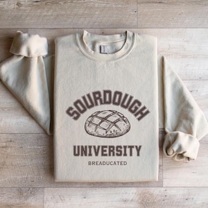 May include: A light beige sweatshirt with the text "SOURDOUGH UNIVERSITY BREADUCATED" and a drawing of a sourdough bread loaf.