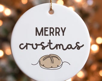 May include: White ceramic Christmas ornament with the words "MERRY crustmas" in dark brown lettering. A cartoon illustration of a bread roll is below the text. The ornament hangs from a gold string.