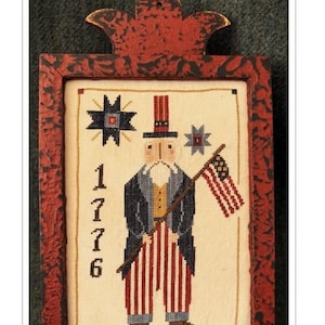 May include: Cross stitch pattern of Uncle Sam in a red, white, and blue outfit holding a flag. The pattern is framed in a red wooden frame with a distressed finish. The finished piece measures 5 inches by 7 inches.