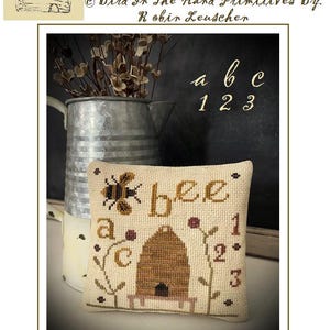 May include: A cross stitch pattern titled "A Bee C Beeskep" by Robin Leuscher. The pattern features a bee, a beehive, and the letters "a", "b", and "c" with the numbers "1", "2", and "3". The finished piece measures 5 1/2 inches by 5 1/2 inches.