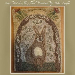 May include: A brown and white needlepoint rug depicting a rabbit in a circle with the words "March Hare" and "©2015 Bird In The Hand Primitives By: Robin Lauschen". The finished piece measures 7 1/2 inches by 5 1/2 inches.