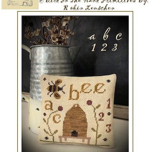 May include: A cross stitch pattern titled "A Bee C Beeskep" by Robin Leuscher. The pattern features a bee, a beehive, and the letters "a", "b", and "c" with the numbers "1", "2", and "3". The finished piece measures 5 1/2 inches by 5 1/2 inches.