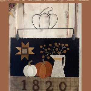 May include: Black wool applique featuring a fall theme with a star, pumpkins, and a pitcher with branches. The bottom section displays the number 1820. Finished piece measures 10 inches by 13 inches.