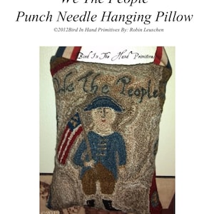 May include: A punch needle pillow with a brown background and a depiction of a person in a blue jacket holding a red, white, and blue flag. The text "We The People" is stitched in brown thread on the pillow. The pillow measures 6 1/2 inches by 8 1/2 inches.