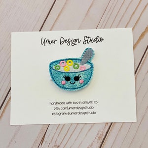 May include: A blue embroidered patch of a bowl of cereal with a spoon. The bowl has a smiling face with eyelashes and pink cheeks. The cereal is yellow, pink, and green. The text "Umer Design Studio" is on the card behind the patch.