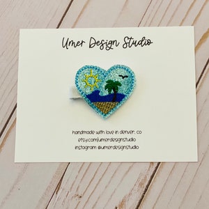 May include: A blue heart-shaped felt hair clip with a sun, palm tree, and ocean scene. The clip is handmade and says "Umer Design Studio" on the card.