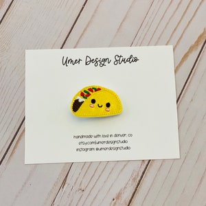 May include: A yellow felt taco with a smiling face, embroidered with green, red, and brown details. The taco is on a white card with the text "Umer Design Studio" and contact information.