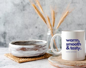 May include: White ceramic mug with the text "warm, smooth & tasty." in blue lettering. The mug is sitting on a wooden coaster and there is a white cloth and a bowl in the background.