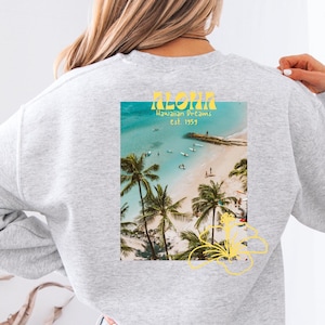 Könnte beinhalten: Grauer Sweatshirt mit einem Grafikdruck einer Strandszene und dem Text "ALOHA" in Gelb. Der Text lautet außerdem "Hawaiian Dreams" und "Est. 1993".