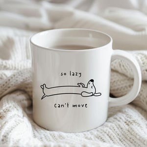 Op de afbeelding: Witte keramische mok met een zwarte illustratie van een teckel en de tekst "so lazy can't move". De mok heeft een gebogen handvat en staat tegen een witte, getextureerde achtergrond.