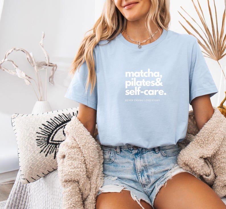 Puede incluir: Una camiseta azul claro con el texto "matcha, pilates & self-care. NEVER ENDING LOVESTORY" impreso en blanco.