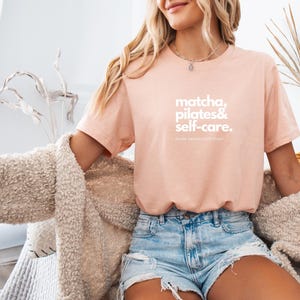Puede incluir: Una camiseta rosa claro con el texto "matcha, pilates & self-care.  Never ending love story" en blanco.