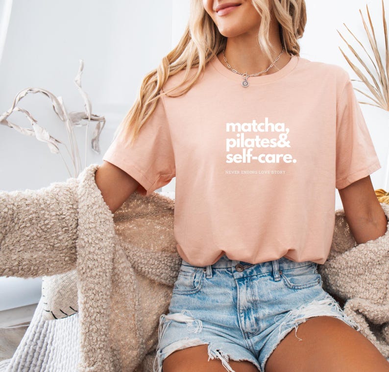 Puede incluir: Una camiseta rosa claro con el texto "matcha, pilates & self-care.  Never ending love story" en blanco.