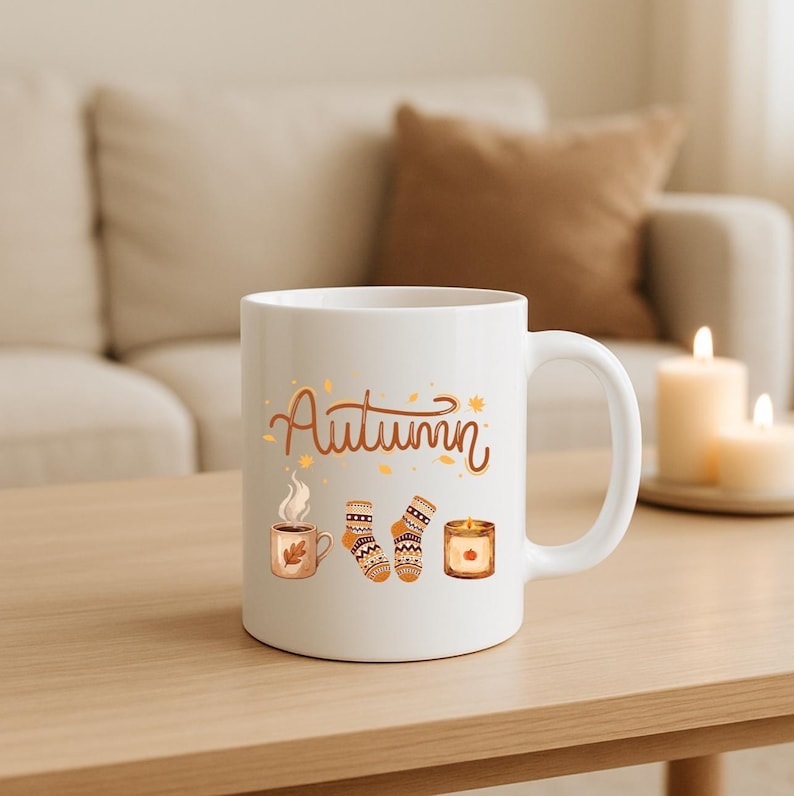 May include: A white ceramic mug featuring the word Autumn in brown script, accompanied by illustrations of a steaming mug, a pair of patterned socks, and a lit candle, all accented with small orange and yellow leaf motifs.