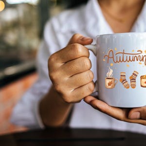 May include: A white ceramic mug featuring the word Autumn in brown script, accompanied by illustrations of a steaming mug, a pair of patterned knit socks, and a lit candle, all rendered in warm orange, brown, and yellow seasonal tones.