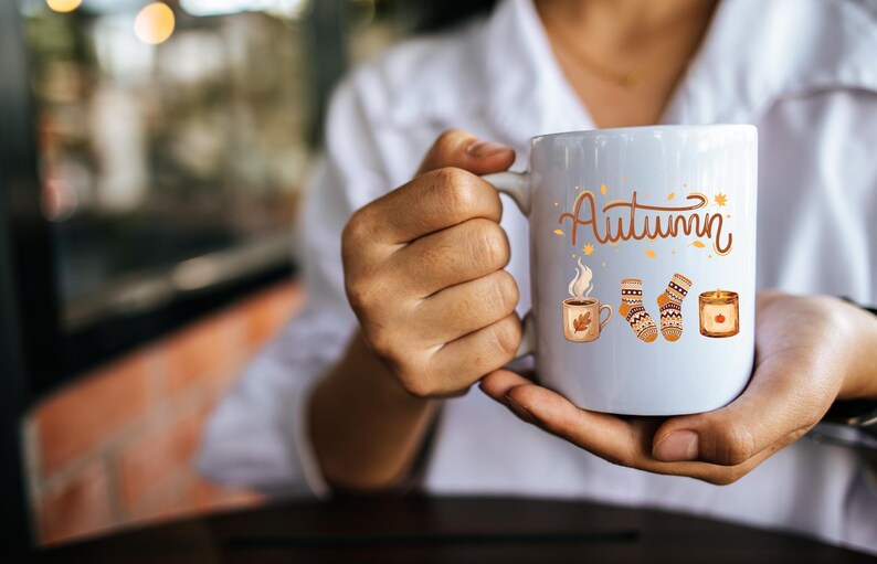 May include: A white ceramic mug featuring the word Autumn in brown script, accompanied by illustrations of a steaming mug, a pair of patterned knit socks, and a lit candle, all rendered in warm orange, brown, and yellow seasonal tones.