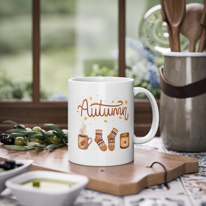 May include: A white ceramic mug featuring the word Autumn in brown script, accompanied by illustrations of a steaming mug, a pair of patterned knit socks, and a lit candle, all surrounded by small scattered orange and brown autumn leaves.