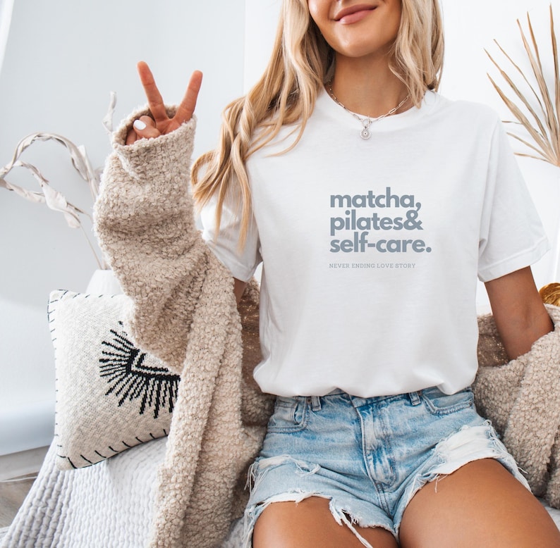 Puede incluir: Una camiseta blanca con el texto "matcha, pilates & self-care. NEVER ENDING LOVE STORY" impreso en gris en la parte delantera.